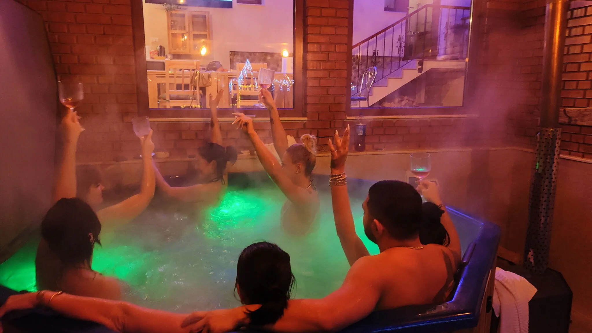 Prieteni în ciubărul tip jacuzzi, cu pahare ridicate, abur și lumină verde de cromoterapie